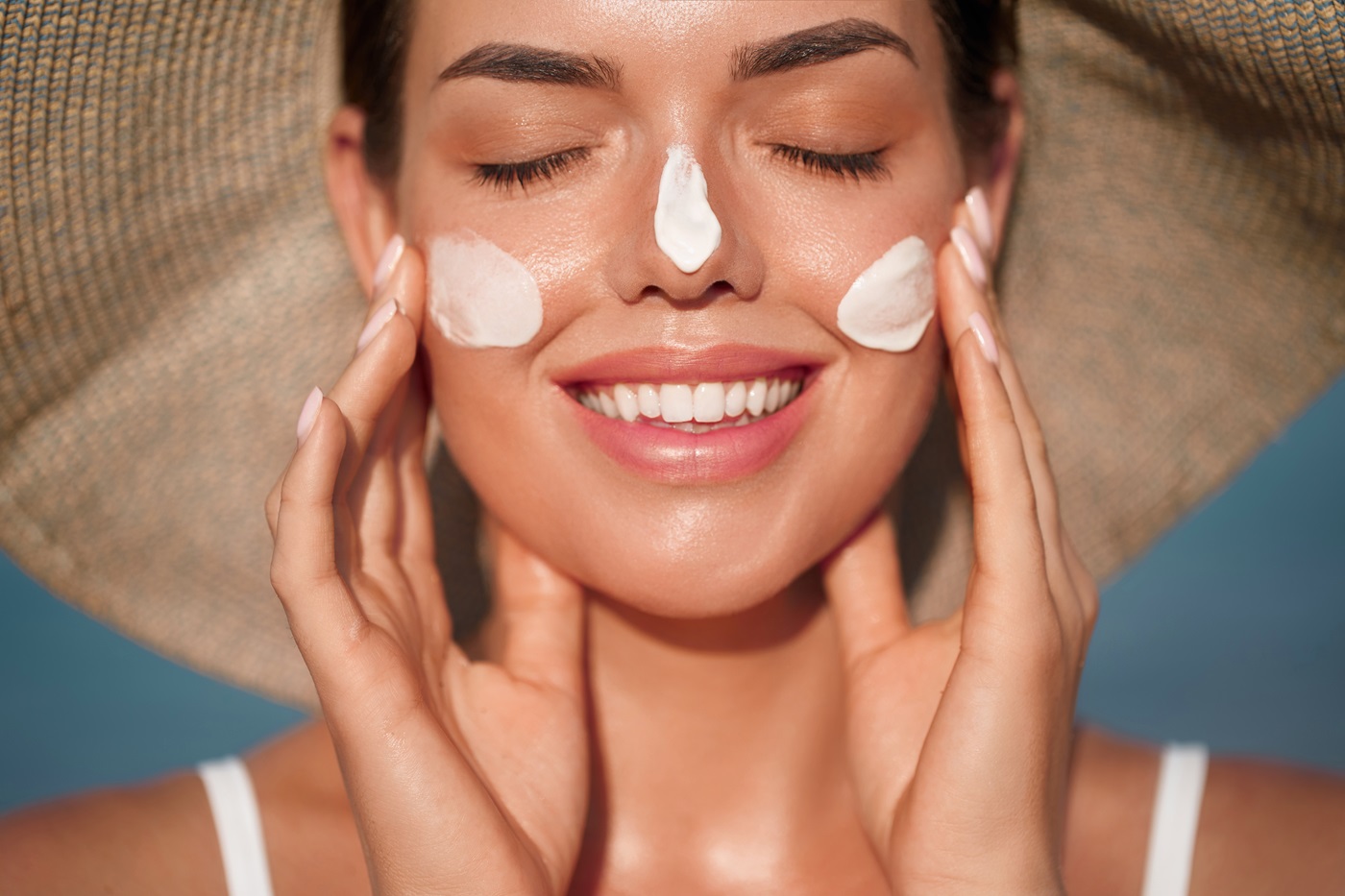 Uśmiechnięta kobieta z zamkniętym dotyka dłońmi policzków, na których ma nałożony najlepszy krem z SPF do twarzy.