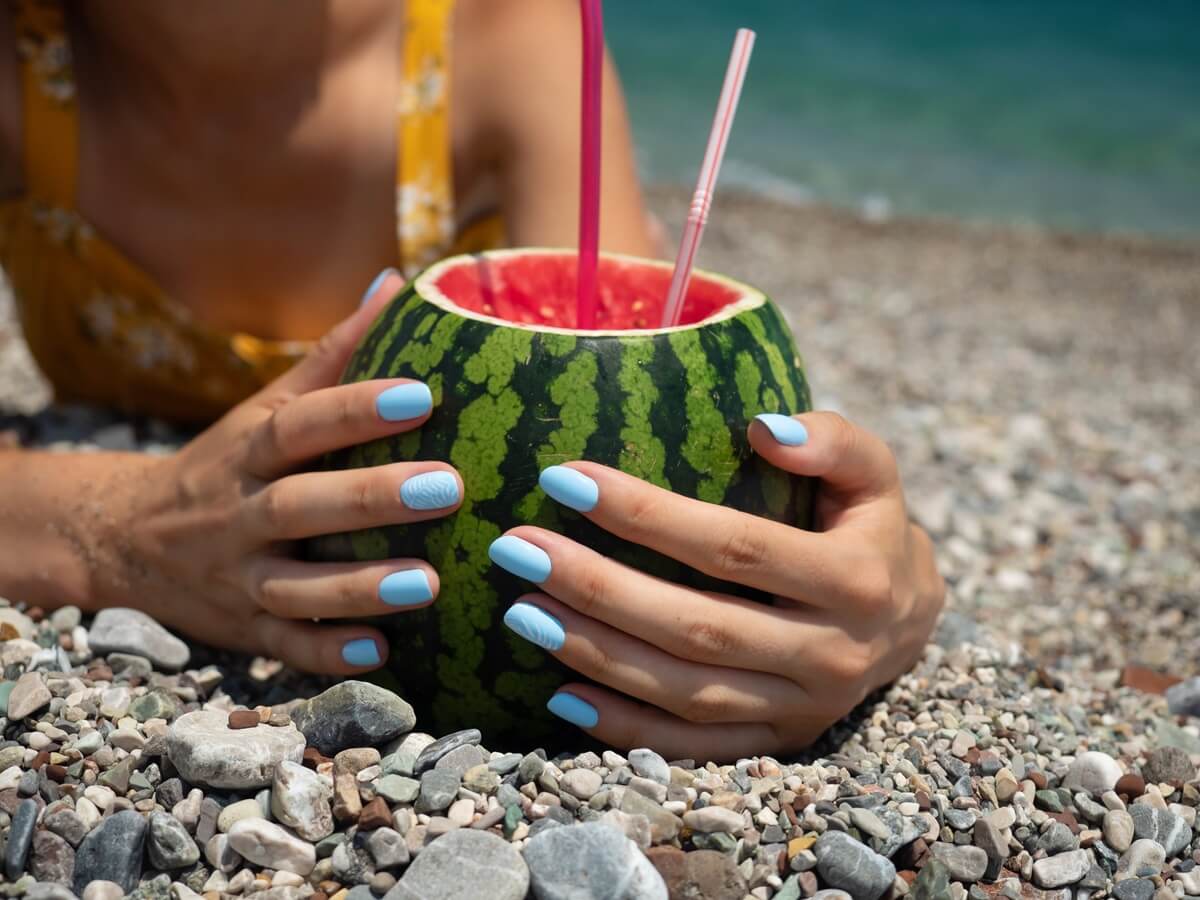Kobiece dłonie z manicure w kolorze sky blue obejmują wydrożonego arbuza ze słomkami w środku na plaży.
