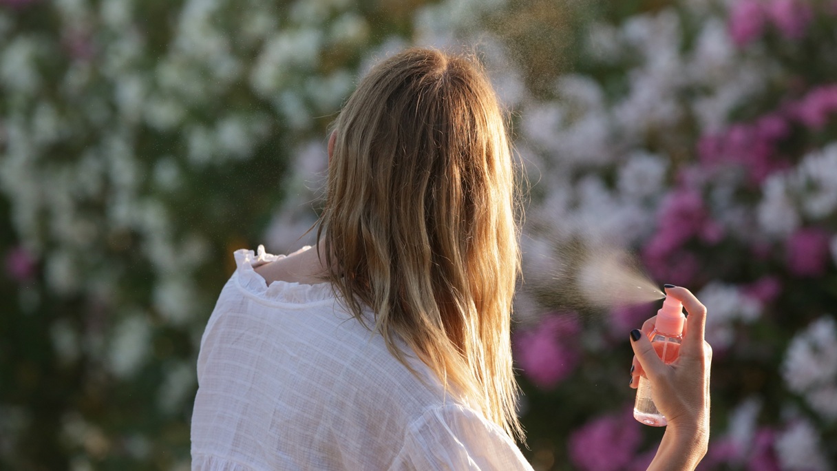 Kobieta z długimi blond włosami rozpyla na włosy perfumy w otoczeniu kwiatów