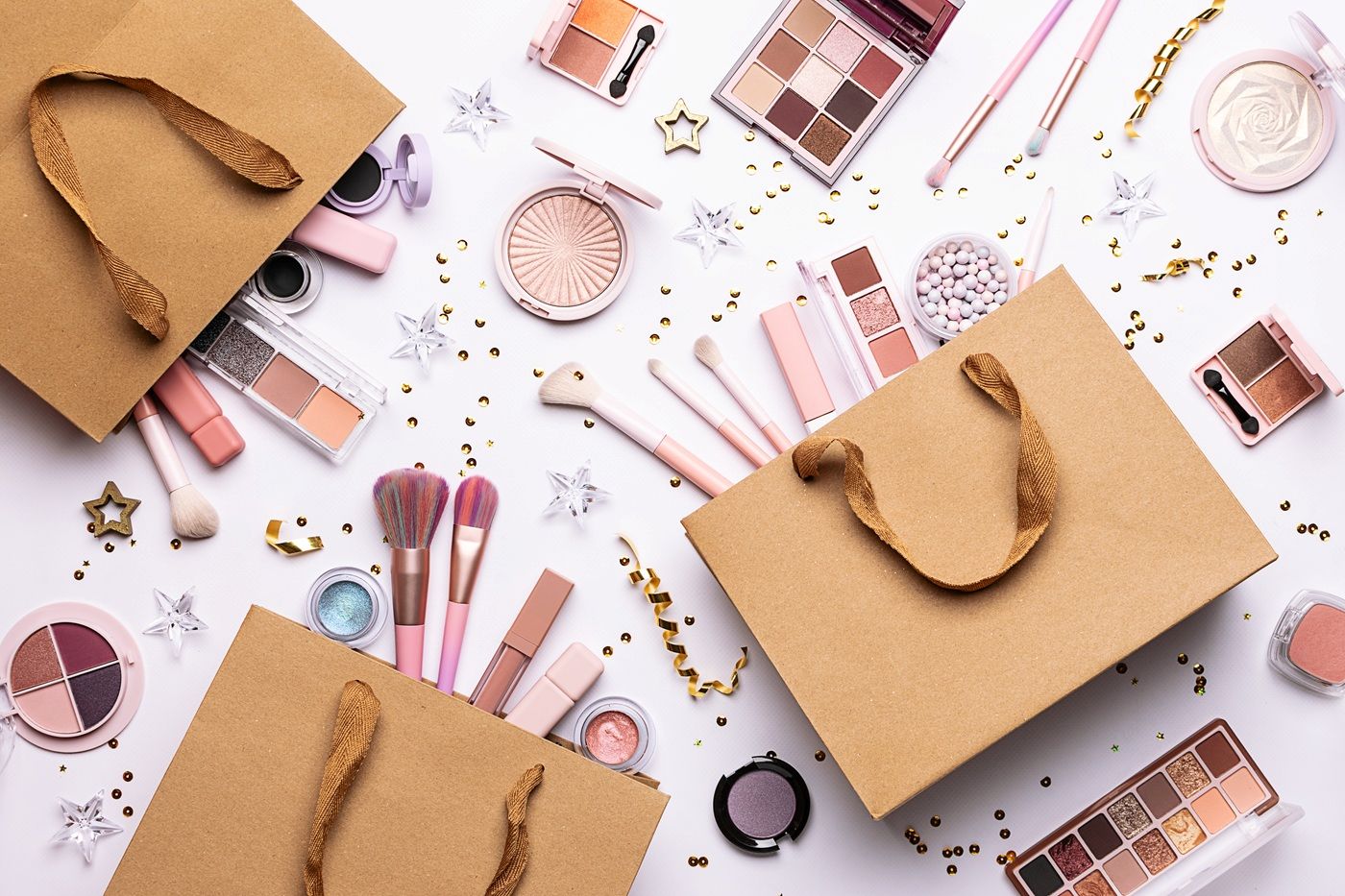 Kosmetyki kolorowe, pędzle do makijażu i ozdobne dodatki rozsypane wokół papierowych torebek prezentowych.
