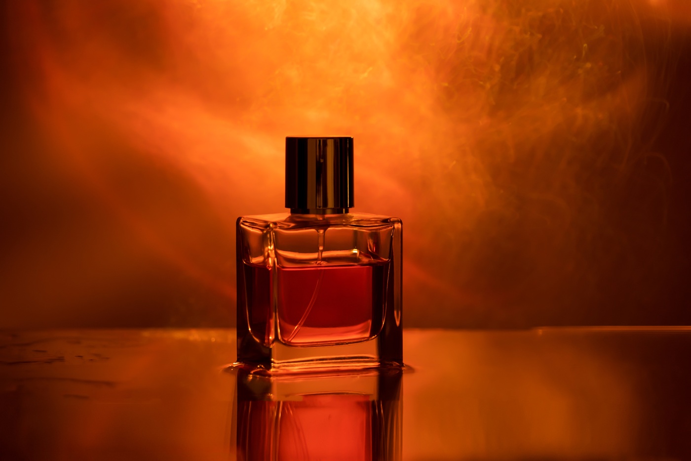 Szklany flakon perfum ustawiony na błyszczącej powierzchni, w ciepłym, pomarańczowo-czerwonym świetle z dymnym tłem.