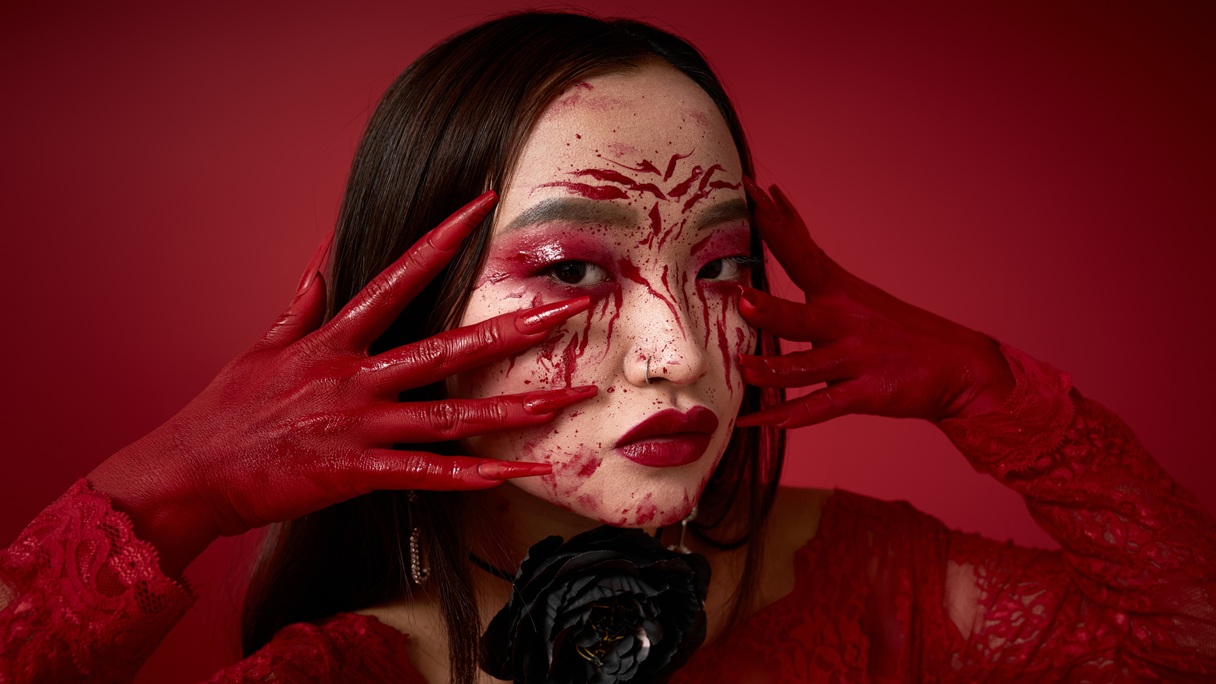 Kobieta w halloweenowym makijażu z imitacją krwawych zadrapań i rozprysków krwi na twarzy, czerwonymi cieniami na powiekach i ciemnoczerwonymi ustami.
