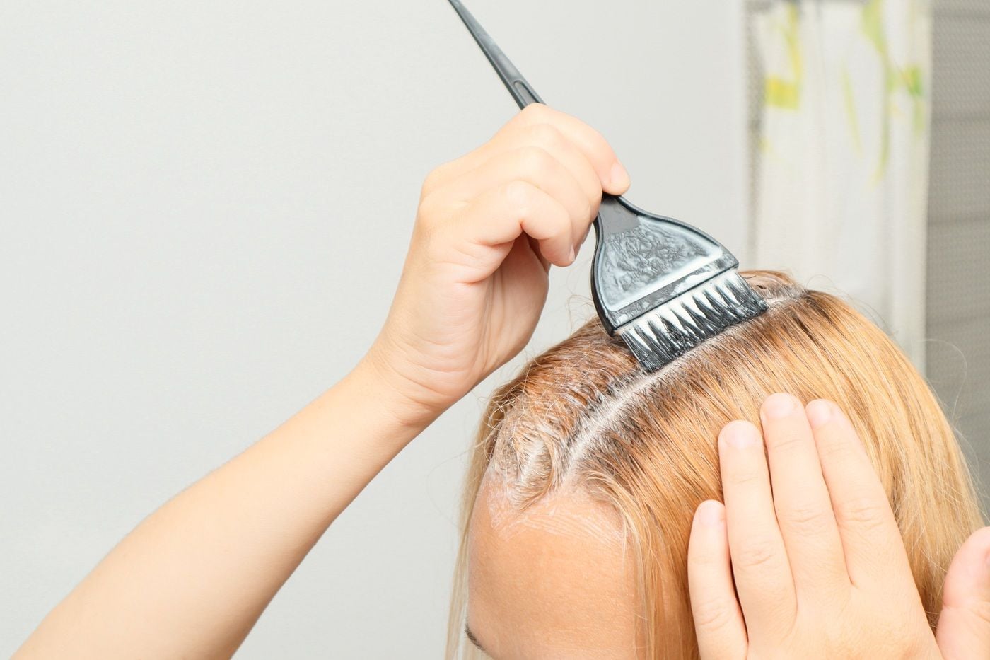 Dłoń nakładająca farbę na blond włosy za pomocą pędzla podczas koloryzacji.