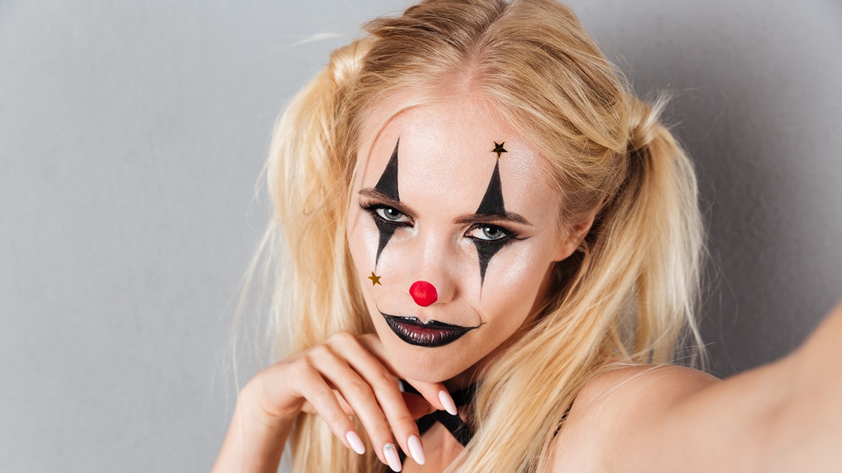 Kobieta w halloweenowym makijażu klauna z czarnymi trójkątami wokół oczu, czerwonym nosem i ciemnymi ustami.