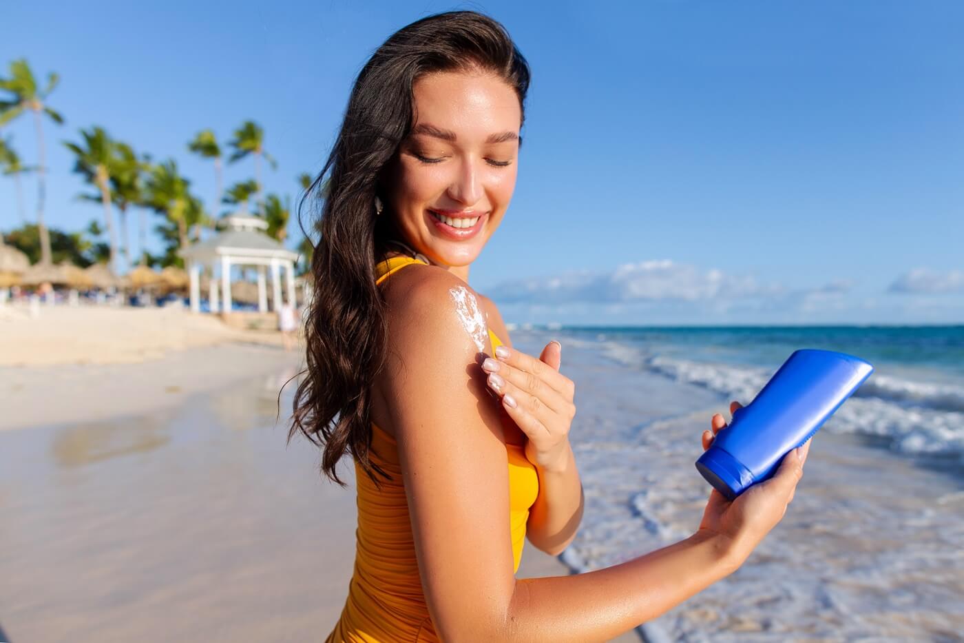 Młoda uśmiechnięta kobieta w żółtym stroju kąpielowym i rozpuszczonych włosach na plaży smaruje ramię kremem z SPF.