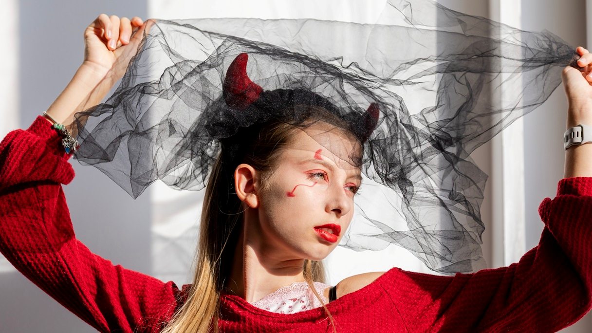Kobieta w makijażu diablicy na Halloween, z czerwonymi ustami, czerwonymi wzorami wokół oczu i opaską z rogami.