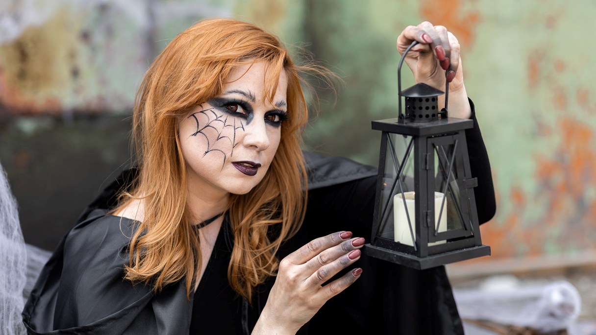 Kobieta w halloweenowym makijażu z czarną pajęczyną na policzku, mocno podkreślonymi oczami i ciemnymi ustami, trzymająca lampion.