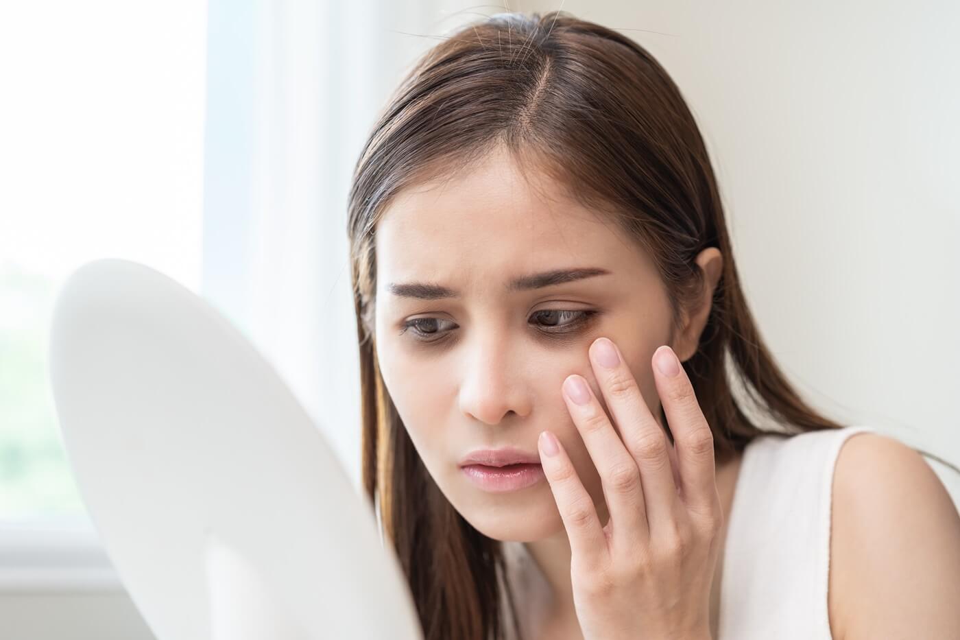 Kobieta z cieniami pod oczami przegląda się w lustrze z zaniepokojoną miną.