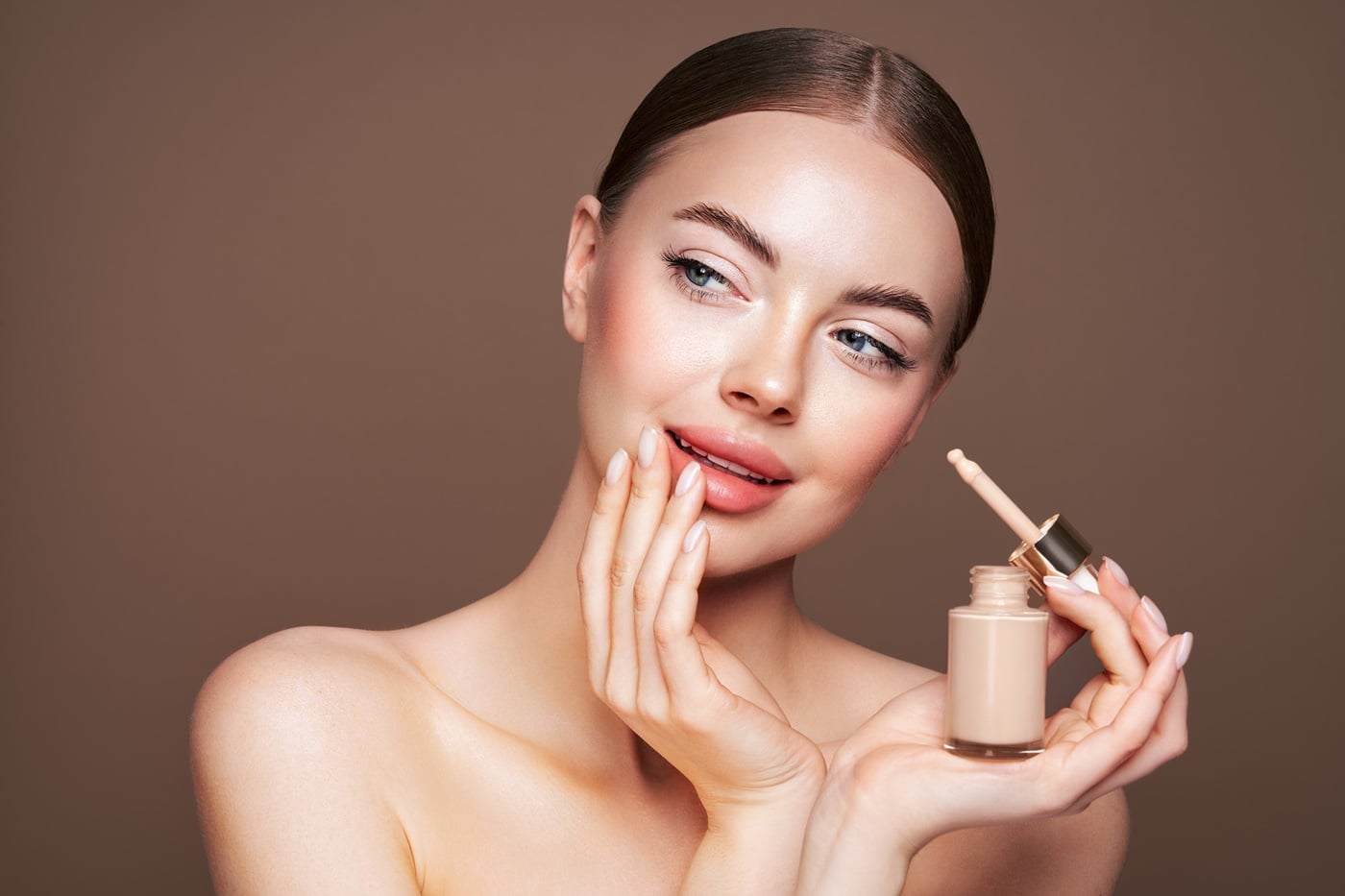 Kobieta trzyma w dłoni buteleczkę płynnego podkładu z aplikatorem na beżowym tle.