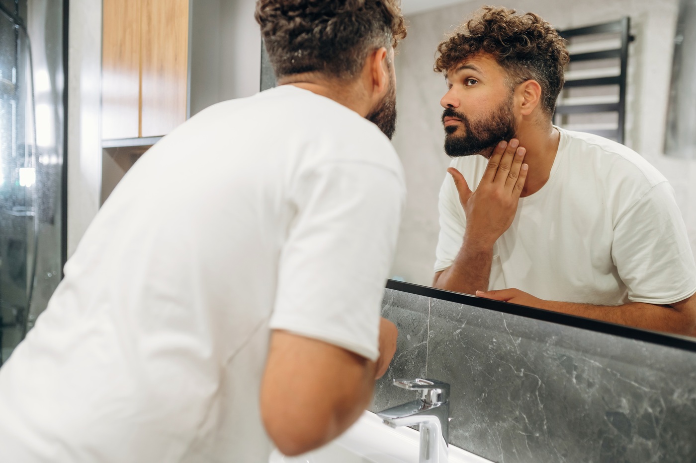 Mężczyzna w białym T-shircie stoi przed lustrem w łazience i ogląda swój zarost