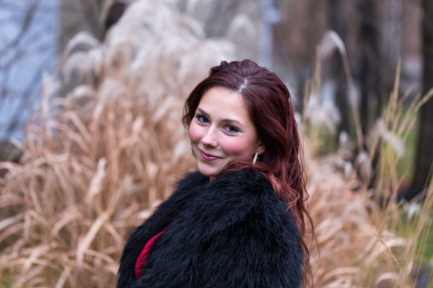 Uśmiechnięta kobieta stoi na tle suchych traw, ubrana w ciemny płaszcz.