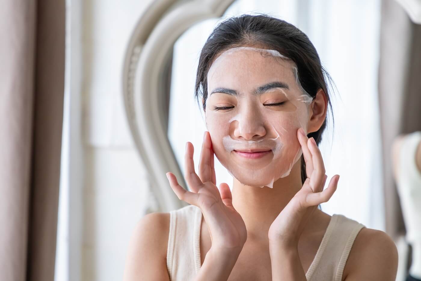 Uśmiechnięta kobieta nakłada na twarz koreańską maseczkę pielęgnacyjną przed lustrem.