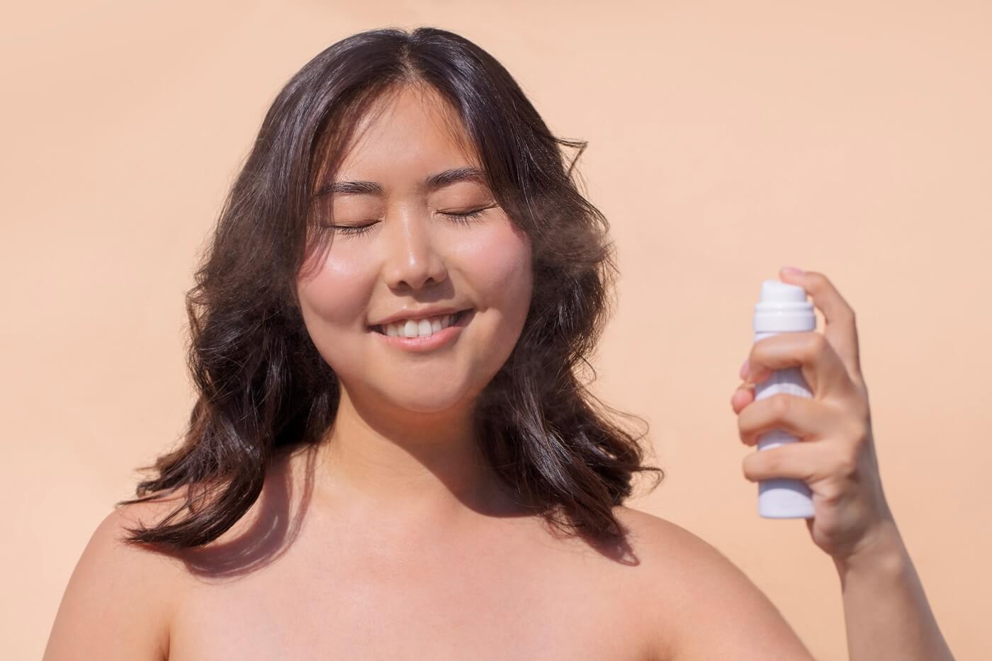 Młoda uśmiechnięta kobieta na beżowym z zamkniętymi oczami używa mgiełki z SPF do twarzy.