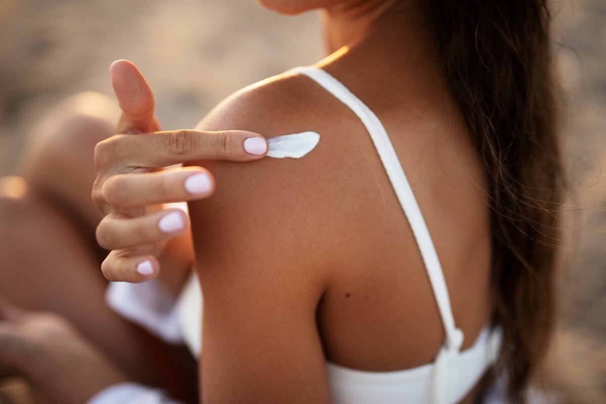 Zbliżenie na kobietę siedzącą na plaży, która rozprowadza biały balsam na opalonym ramieniu.