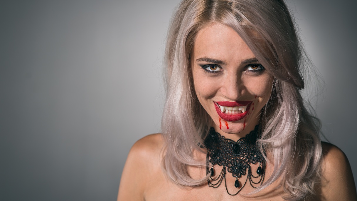 Kobieta w makijażu wampirzycy na Halloween, z czerwonymi ustami, sztuczną krwią spływającą z kącików ust i mocno podkreślonymi oczami.