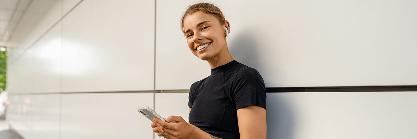 Młoda kobieta uśmiechająca się do kadru, w ręku trzyma telefon